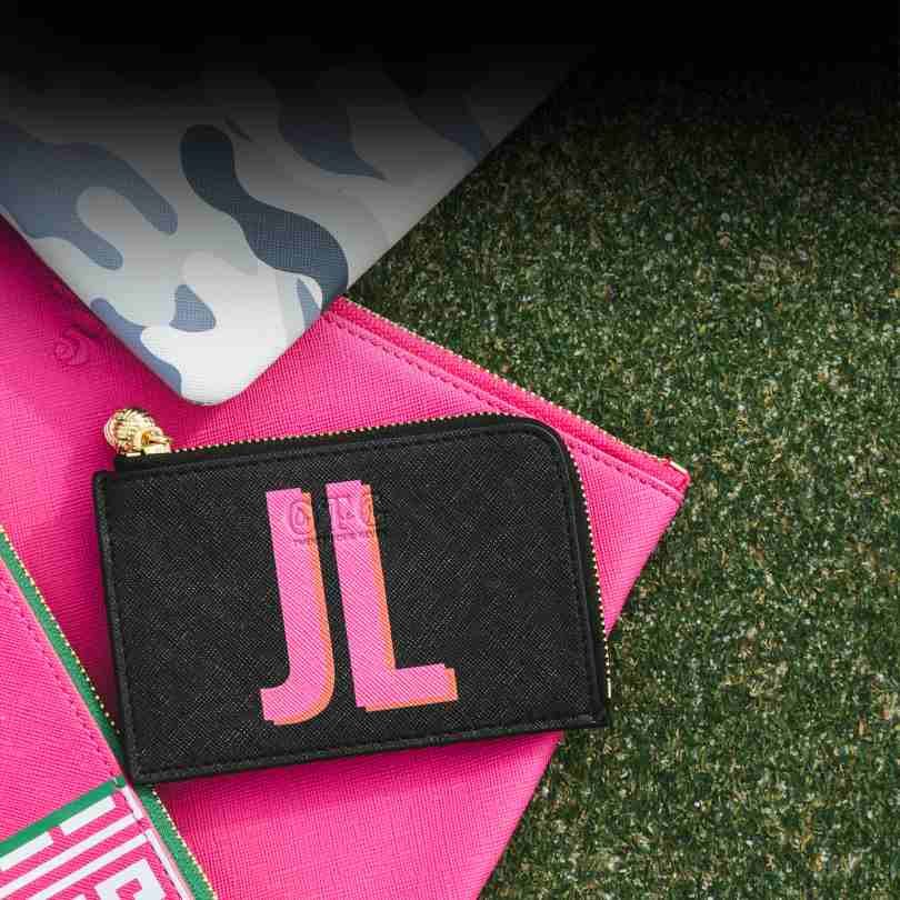 A black card case personalized with initials "JL" in Pink and Orange writing. The card case sits on green grass and hot pink bags