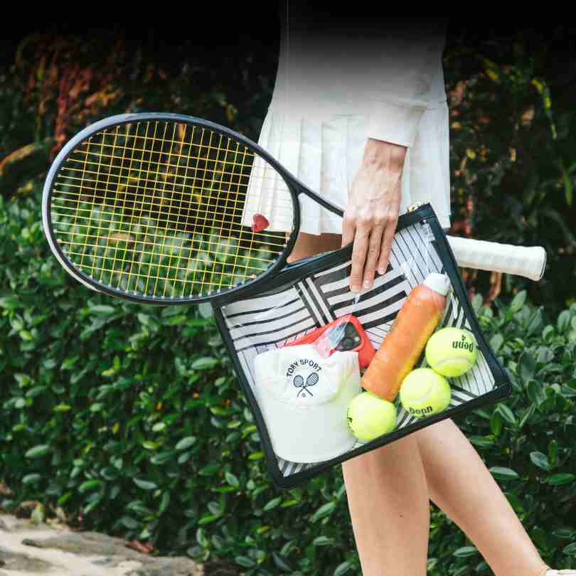 A women carrying a clear black bag with tennis balls, sunscreen and a hat. She has a tennis skirt on and holding a tennis racquet