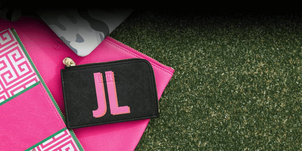 A black card case personalized with initials "JL" in Pink and Orange writing. The card case sits on green grass and hot pink bags