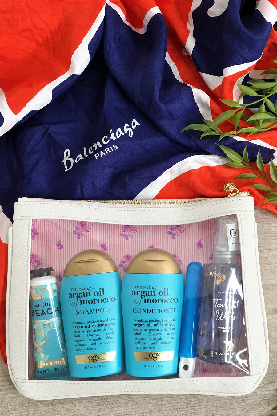 White floral #4 Meeghan Nudie clear-window pouch packed with travel-size shampoo, conditioner and sunscreen, styled on a silk scarf